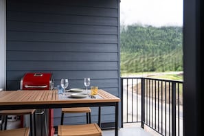 A wooden table with a red grill, two wine glasses, and plates on a balcony with a view of a forested area. - Summit Views by Revelstoke Vacations (Revelstoke)