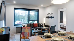 Ein modernes Wohnzimmer mit einer Couch, einem Esstisch gedeckt mit Tellern und Gläsern, einem großen Fenster und einer Wandkunst mit Berglandschaft.
