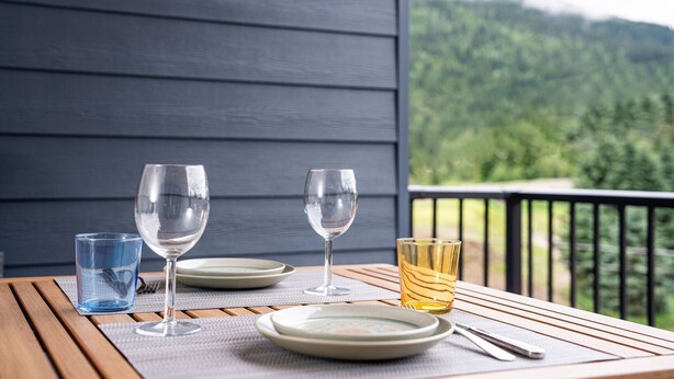 Ein Tisch mit Tellern, Gläsern und Besteck auf einer Holzterrasse mit Blick auf einen bewaldeten Gebirgszug.