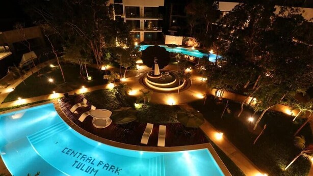 A nighttime view of a pool area with a central fountain, surrounded by well-lit pathways and greenery.