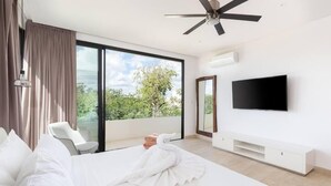 A modern hotel room with a large bed, a flat-screen TV, a ceiling fan, and a balcony with a view of greenery.