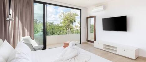 A modern hotel room with a large bed, a flat-screen TV, a ceiling fan, and a balcony with a view of greenery.