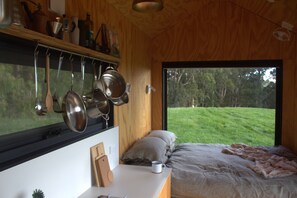 A bedroom with a large window, a bed, a bedside table with a cup, and kitchen utensils hanging above.