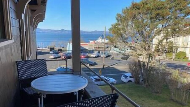 A balcony with two chairs and a table, overlooking a parking area and a body of water with mountains in the distance. - The Historic Downtown Astoria Flats (Astoria)