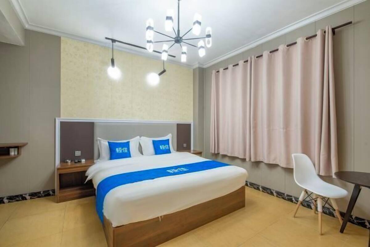 A hotel room with a bed, bedside tables, a desk, a chair, a ceiling light fixture, and a window with curtains.