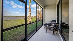 A balcony with large windows, wicker furniture, and a view of palm trees and a body of water.