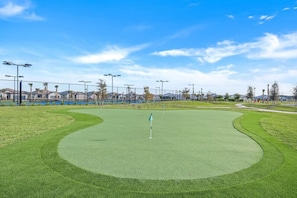 Un campo de golf con un putting green, rodeado por una valla, y casas al fondo.