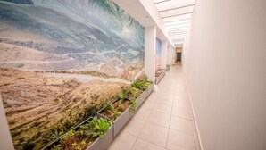 Un couloir orné d’une grande fresque représentant un paysage, doté d’un sol carrelé et de jardinières remplies de plantes vertes.