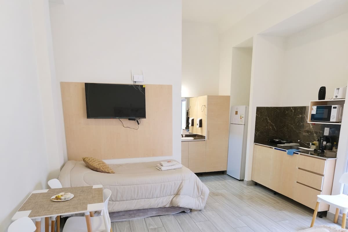 A modern hotel room with a dining table, two beds, a TV, a kitchen area, and a flat-screen TV mounted on the wall.