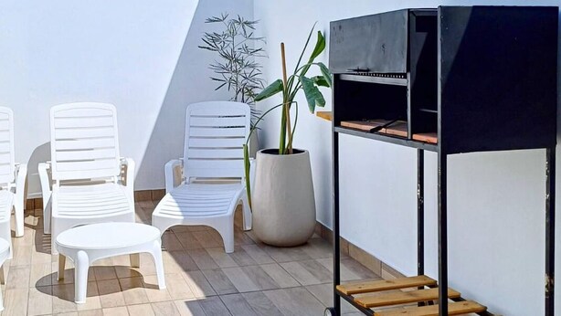 A patio with white plastic chairs, a small round table, a potted plant, and a black grill with a wooden shelf.