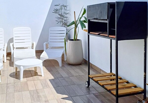 A patio with white plastic chairs, a small round table, a potted plant, and a black grill with a wooden shelf.