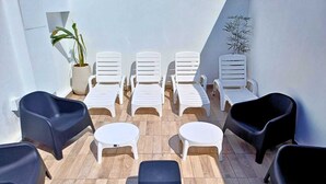 Un espace extérieur lumineux, aménagé avec des chaises longues blanches et des fauteuils noirs, entouré de plantes en pot et doté d’un plancher en bois.