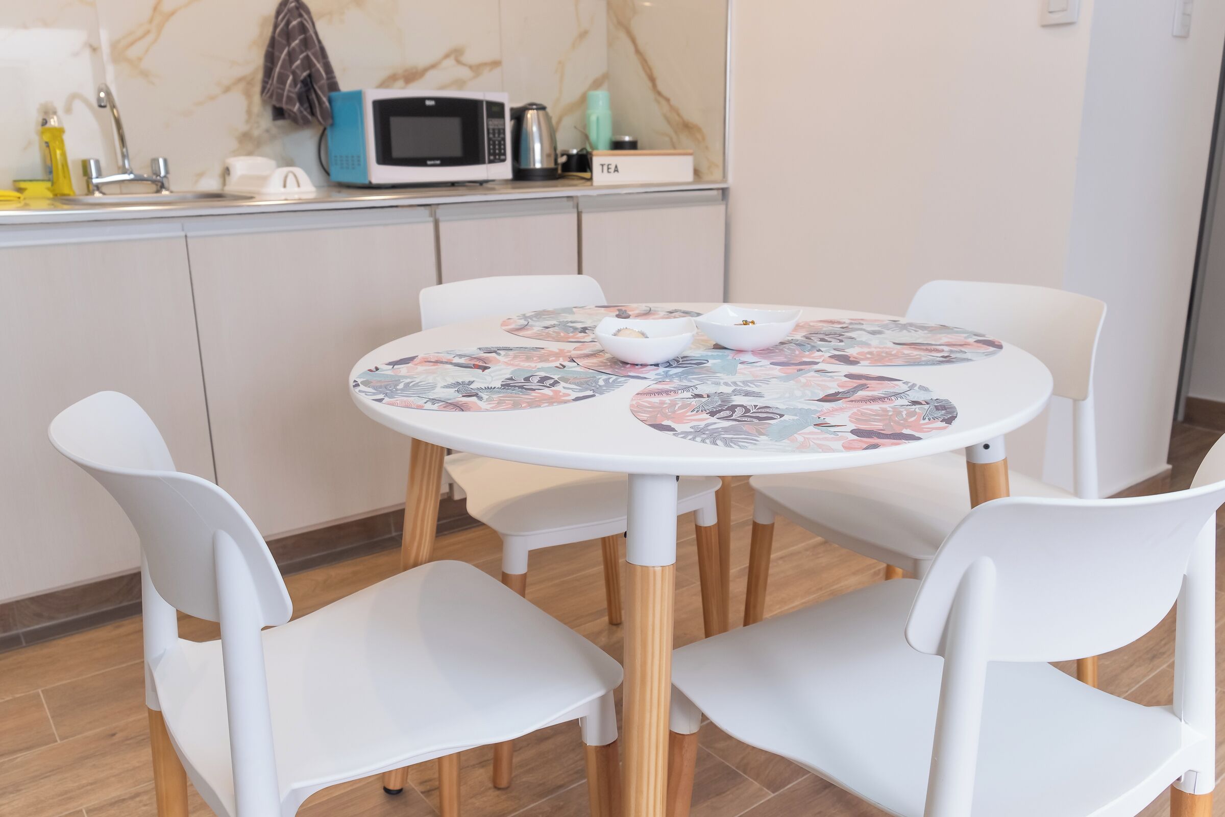 A modern kitchen with white cabinets, a microwave, and a kettle. There is a small table with a floral tablecloth and white chairs.