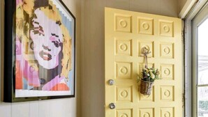 A yellow door with a decorative basket hanging on it, a framed artwork on the wall, and a wooden floor. - Sunny's Haven in Little Italy (Omaha)