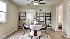 A dining area with a round table, two chairs, a ceiling fan, and shelves with decorative items. - Sunny's Haven in Little Italy (Omaha)