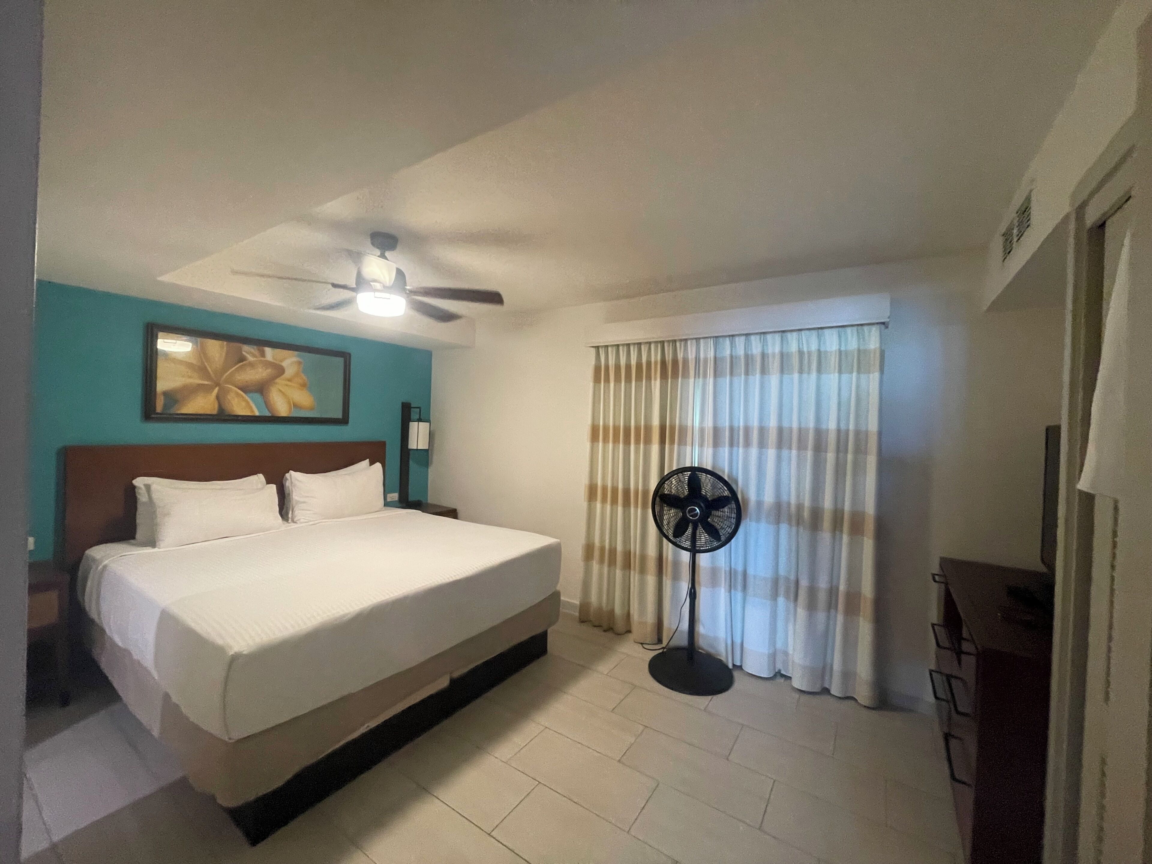 Une chambre d’hôtel avec un lit, un ventilateur de plafond, un ventilateur sur pied et une fenêtre avec des rideaux.