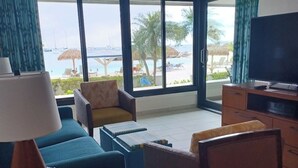 A hotel room with a large window offering a view of the sea and beach. There is a ceiling fan, a TV, and a seating area with a sofa and chairs. - Amazing Beachfront Condo in Simpson Bay -Walk Out Beach/Pool (Philipsburg)