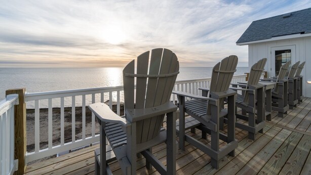 A deck with Adirondack chairs facing a body of water, a white building, and a wooden deck. - Soundfront 10BR with Pool & Hot Tub- Perfect for Large Groups ★★★★★ (Avon)