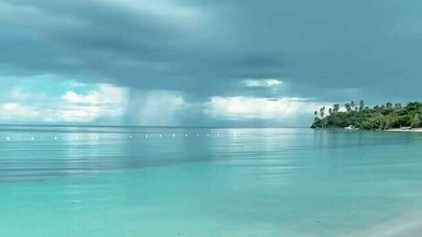 Una playa con agua turquesa cristalina, una orilla arenosa y una vista distante de una isla boscosa bajo un cielo nublado.