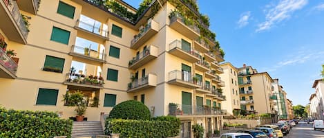 A multi-story residential building with balconies, a gated entrance, and parked cars along the street.