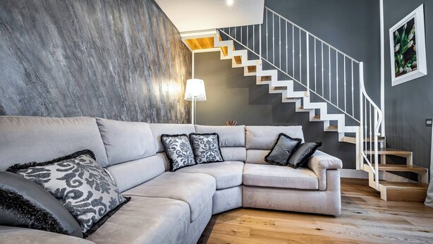 Ein modernes Wohnzimmer mit einem grauen Ecksofa, einer Holztreppe und einer Wandleuchte.