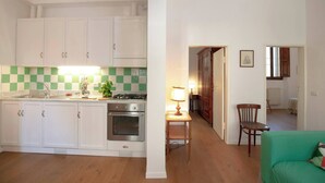 Una cocina compacta con armarios blancos, una estufa y un lavamanos. En la zona de estar contigua, hay una mesa pequeña con una lámpara y una silla.