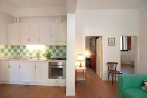 Una cucina compatta con mobili bianchi, un fornello e un lavello. Nell'area soggiorno adiacente, si trova un piccolo tavolo con una lampada e una sedia.