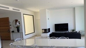 A modern living room with a large white dining table, white chairs, a black sofa, and a flat-screen TV. - Villa Napoleon, vue mer Exceptionnelle, Grande Piscine à Debordement (Pietrosella)