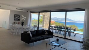 A modern living room with a black leather sofa, dining table, and chairs. Large windows offer a view of the sea and mountains. - Villa Rivoli - Domaine Napoleon Corsica vue mer Exceptionnelle Grande Piscine (Pietrosella)
