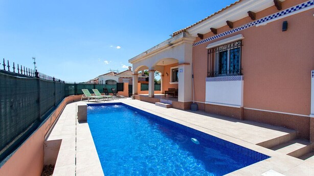 Una piscina con plataforma de baldosas, un área de estar techada y un edificio con fachada y ventanas de cerámica.