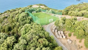 An aerial view of a coastal area with a soccer field surrounded by dense greenery and a rocky shoreline. - Two-room apartment of approximately 42 m2 with air conditioning composed of a living room with TV, foldaway bed and single sofa bed, kitchenette equipped with electric hobs and fridge.Bedroom with double bed, bathroom with shower and hairdryer, shaded ve (Teulada)