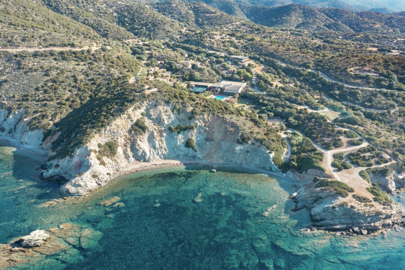 An aerial view of a coastal property with clear turquoise waters, rocky cliffs, and a building with a swimming pool.