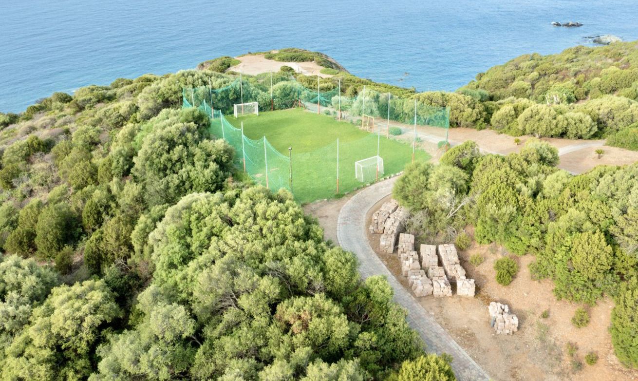 Vue aérienne d’une zone côtière avec un terrain de football entouré d’une végétation luxuriante et bordé d’une côte rocheuse.