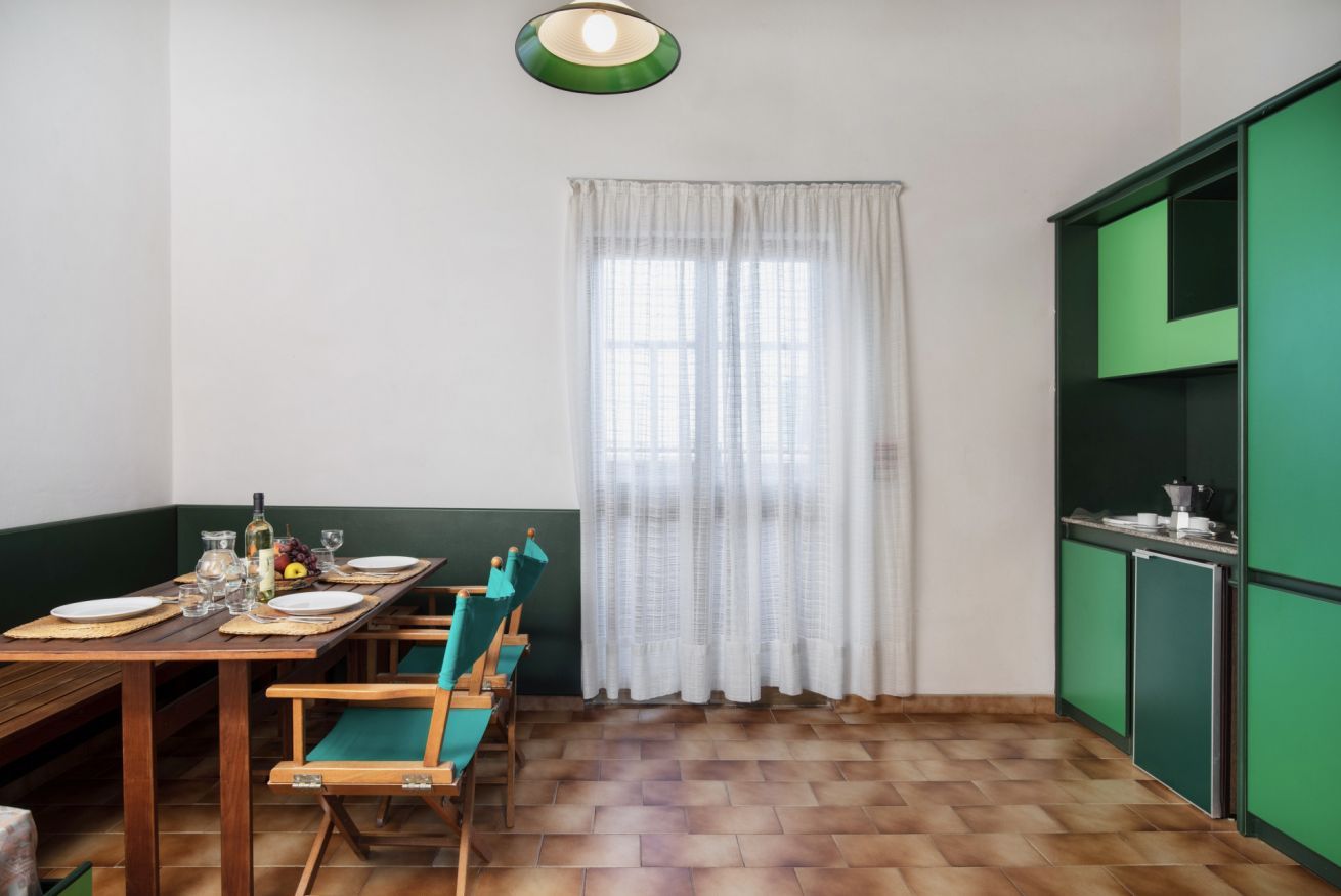Un coin repas avec une table en bois dressée pour quatre personnes, des chaises vertes et une fenêtre ornée de rideaux blancs.
