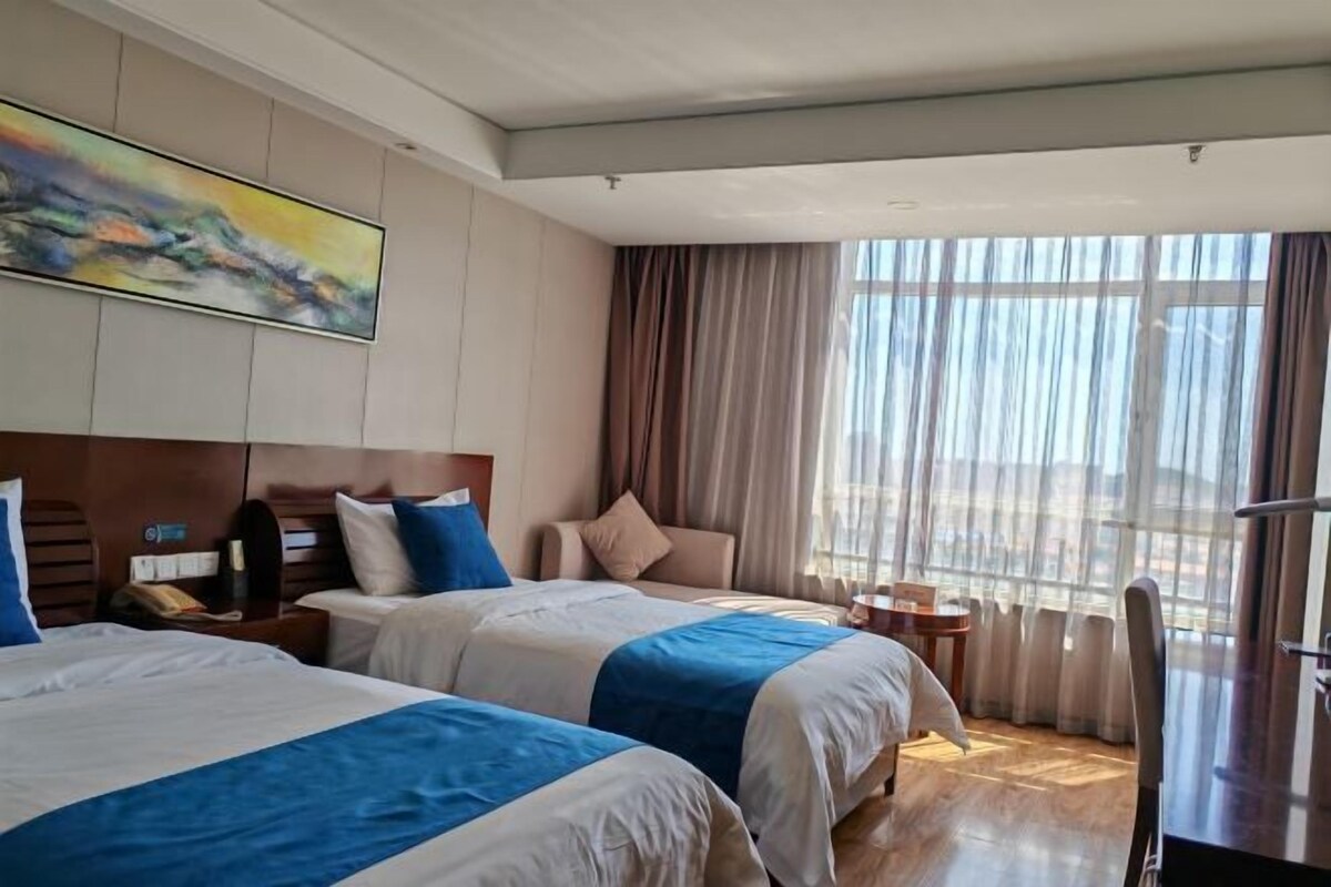 A hotel room with two beds, a large window with curtains, a painting on the wall, and a small table with a chair.