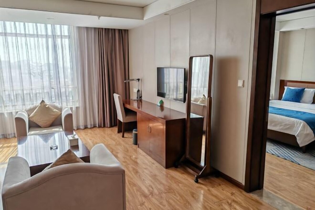 A hotel room with a wooden floor, a desk with a chair, a bed with blue bedding, a TV, and a large mirror.