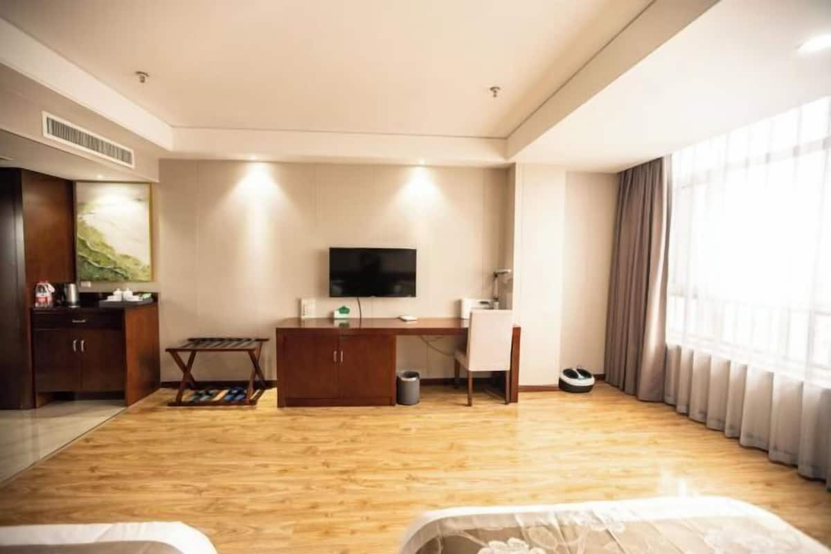 A hotel room with a wooden floor, a desk with a chair, a TV mounted on the wall, a bed, and a window with curtains.