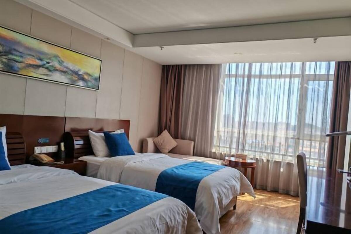 A hotel room with two beds, a large window with curtains, a painting on the wall, and a small table with a chair.