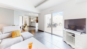 A modern living room with a sofa, coffee table, and TV stand. There is a dining area with chairs and a table, and a kitchen in the background.