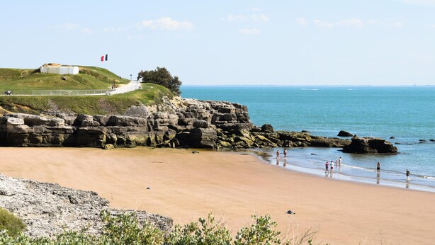 A coastal landscape with a sandy beach, rocky cliffs, and a grassy hill with a building and flag.
