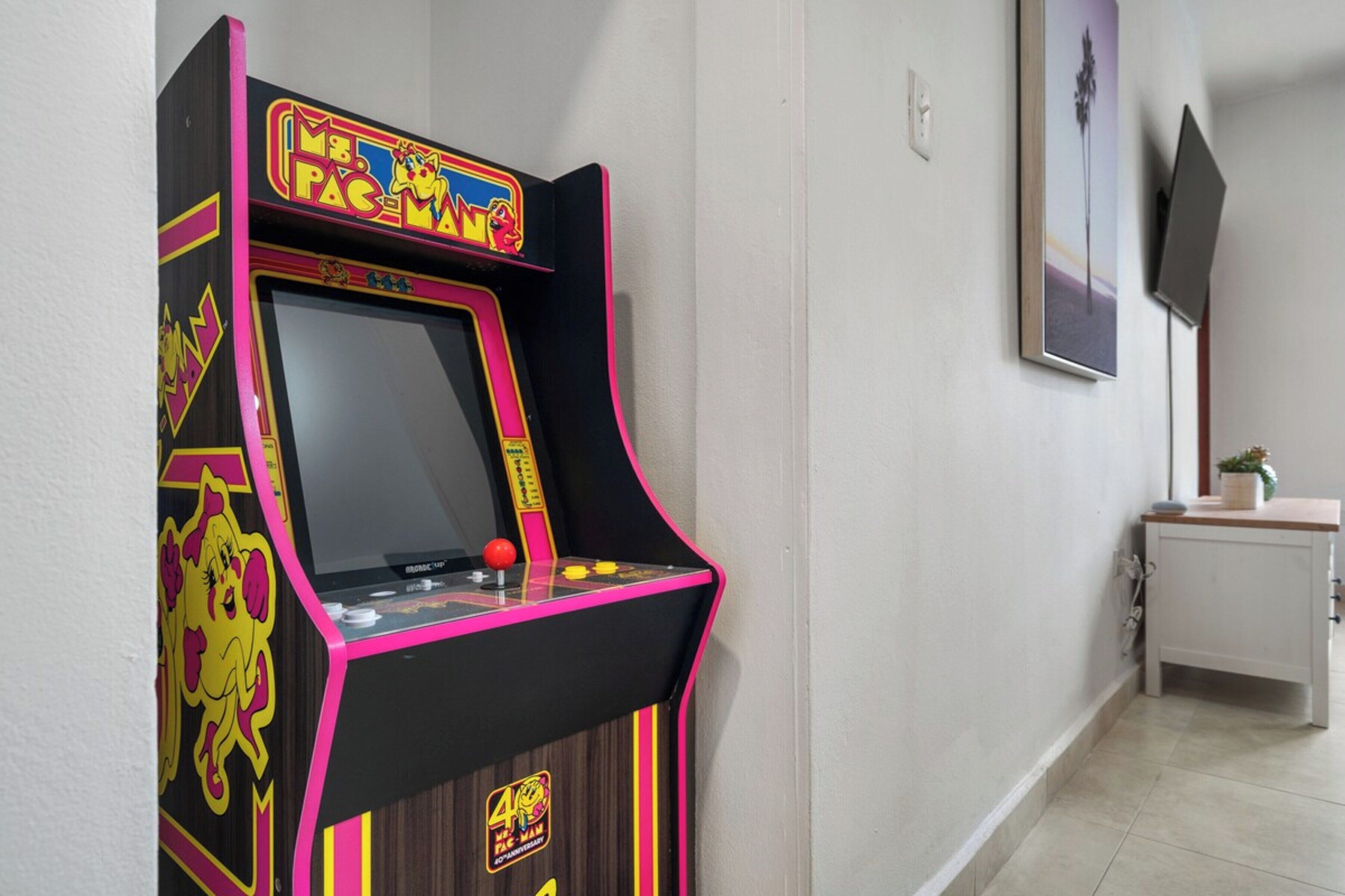 Una macchina arcade Pac-Man in un corridoio con un muro bianco e un quadro incorniciato sopra.