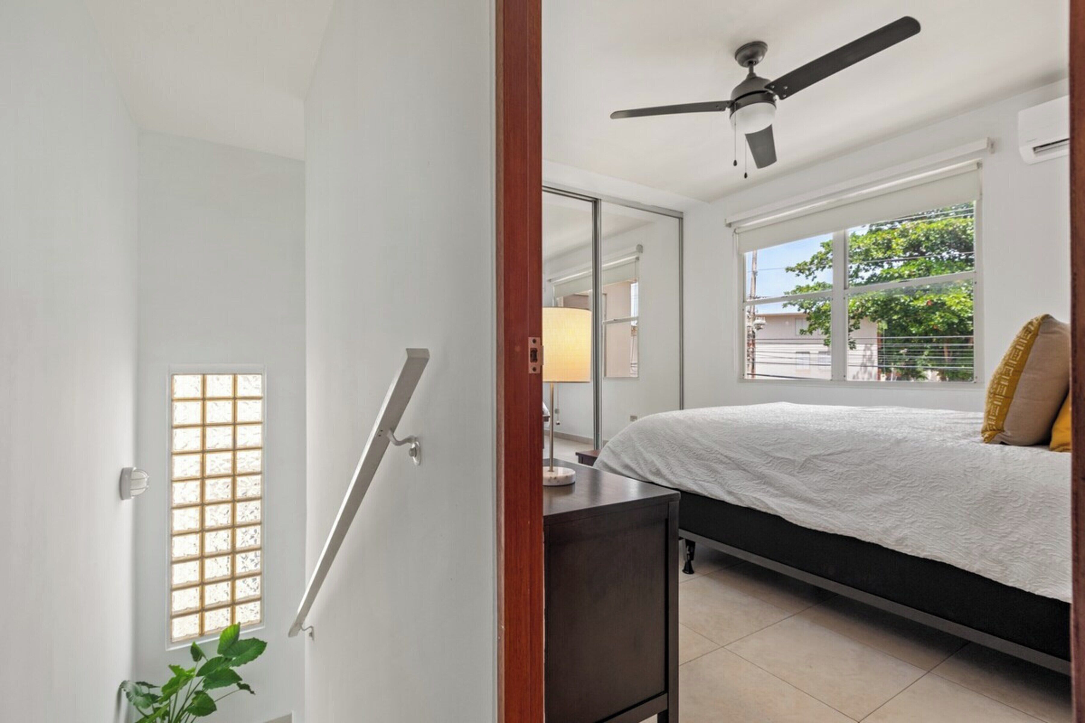 Ein Schlafzimmer mit Bett, Deckenventilator, Fenster mit Außenblick und Tür mit Milchglasfenster.