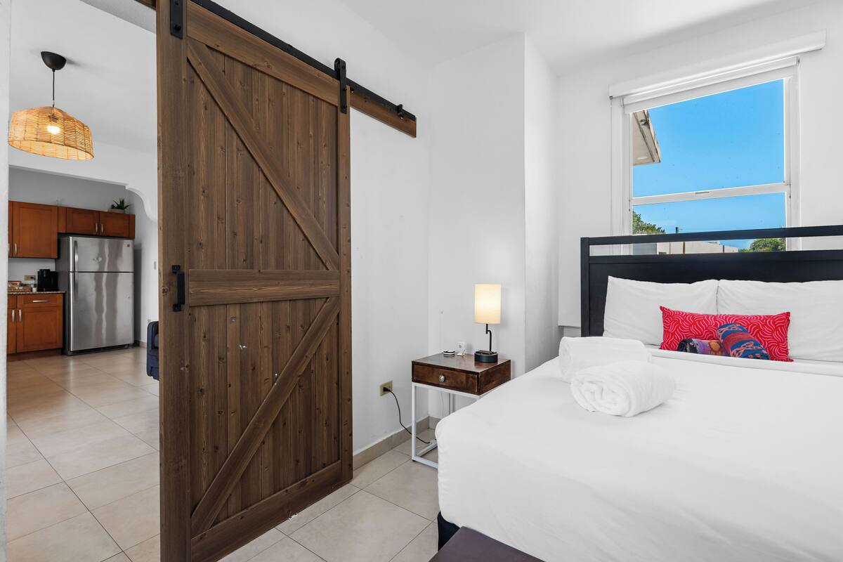 Hotelkamer met een bed, nachtkastje, lamp, houten deur, keuken en raam met uitzicht.