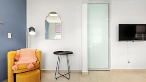 Una habitación con un sillón amarillo, una mesa redonda pequeña, un espejo y un televisor montado en la pared.
