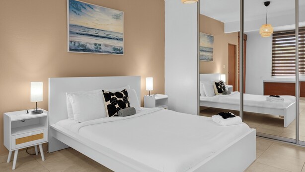 Una habitación de hotel moderna con una cama grande, mesitas de noche con lámparas, un cuadro en la pared y un espejo grande que refleja la habitación.