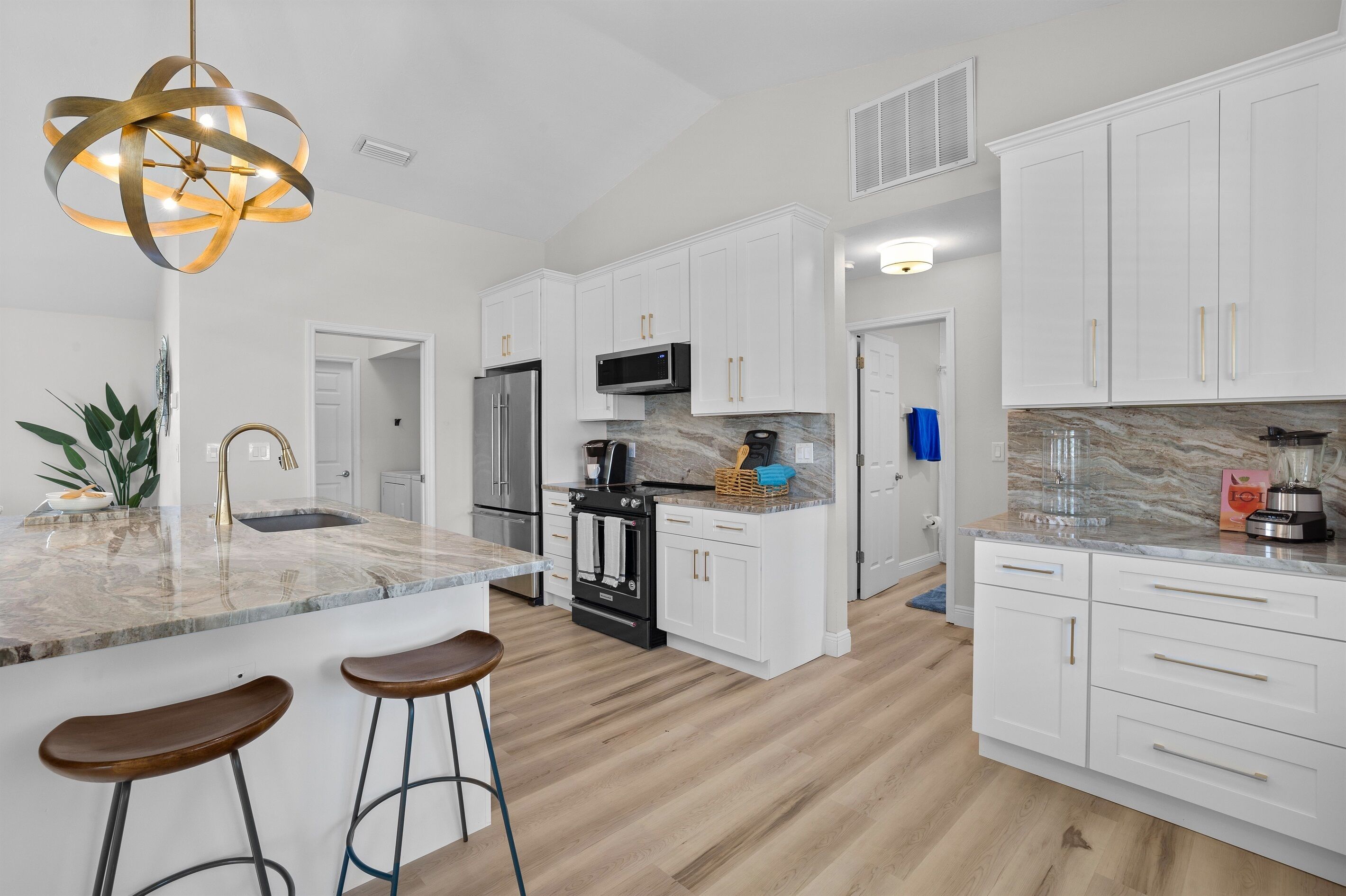 A modern kitchen with white cabinets, a stainless steel refrigerator, a microwave, and a built-in oven. There are two wooden bar stools and a marble countertop.