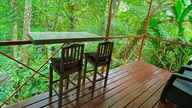 An outdoor wooden deck with a table and chairs, surrounded by lush greenery. - Cabañas El Bosque del Arenal (Peñas Blancas)