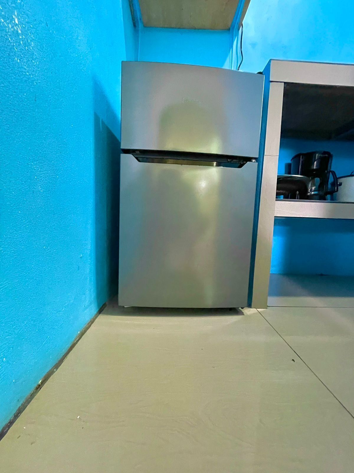 Una cocina compacta con un refrigerador de acero inoxidable, una cafetera y una estantería.