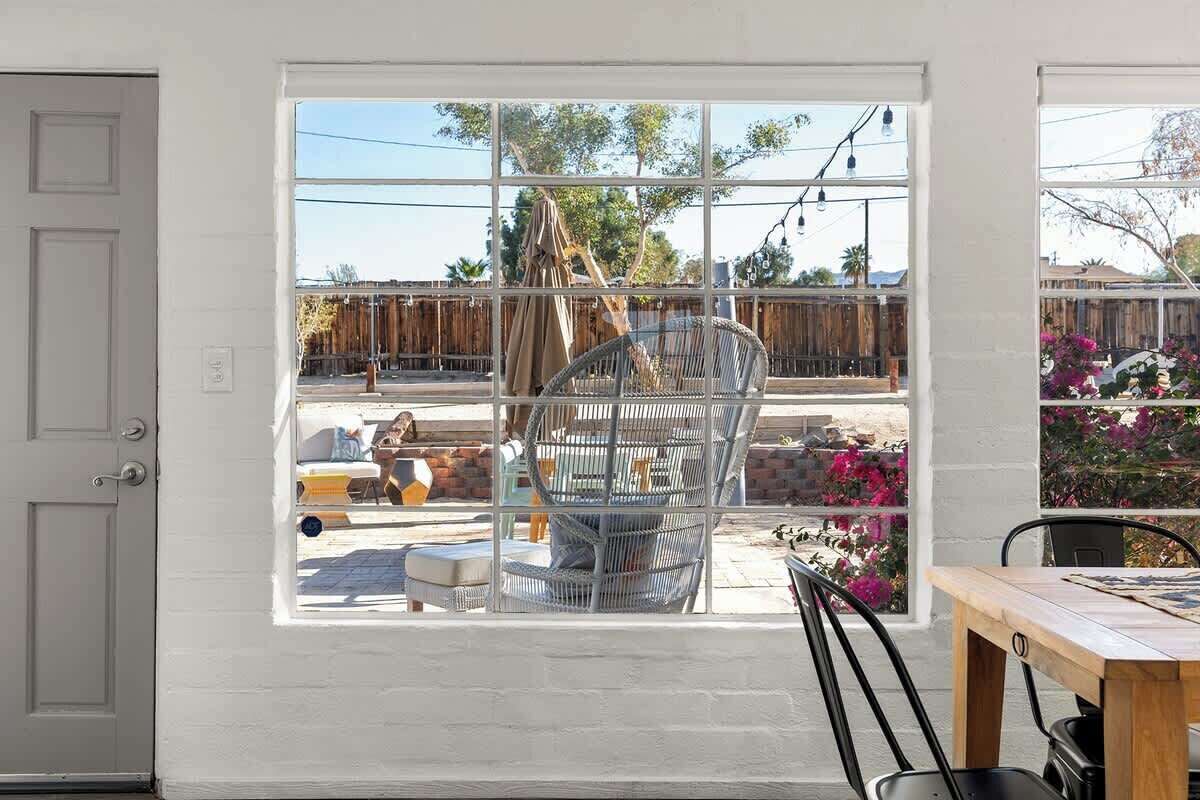 Ein Zimmer mit großem Fenster, einem Korbstuhl, einem Holztisch und einer Tür.