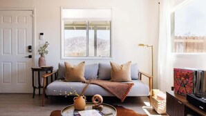 A living room with a sofa, a coffee table, a bookshelf, a window with a view, and a door. - The Creosote Cottage| A Luxury Desert Escape (Landers)